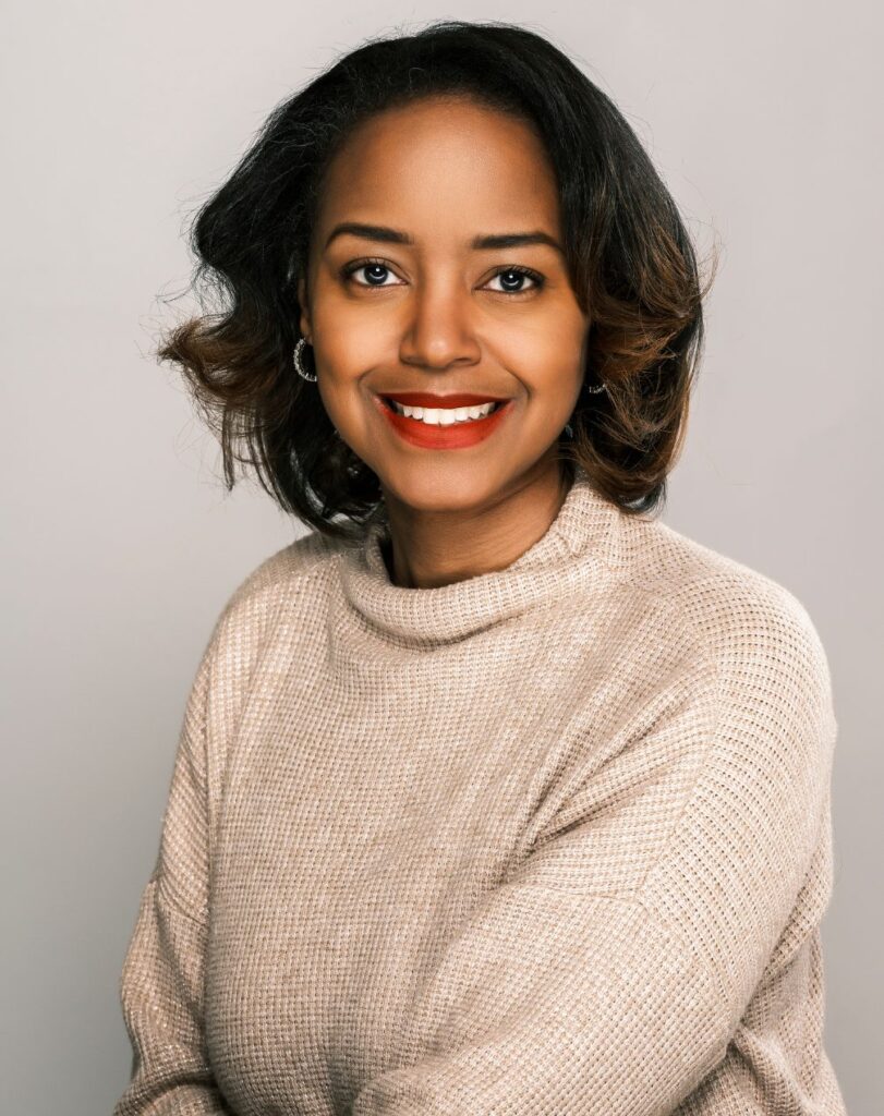 Joy A. Johnson, a young or middle-aged Black woman with short shoulder-length dark hair. She wears a cream turtleneck, hoop earrings, and red lipstick.