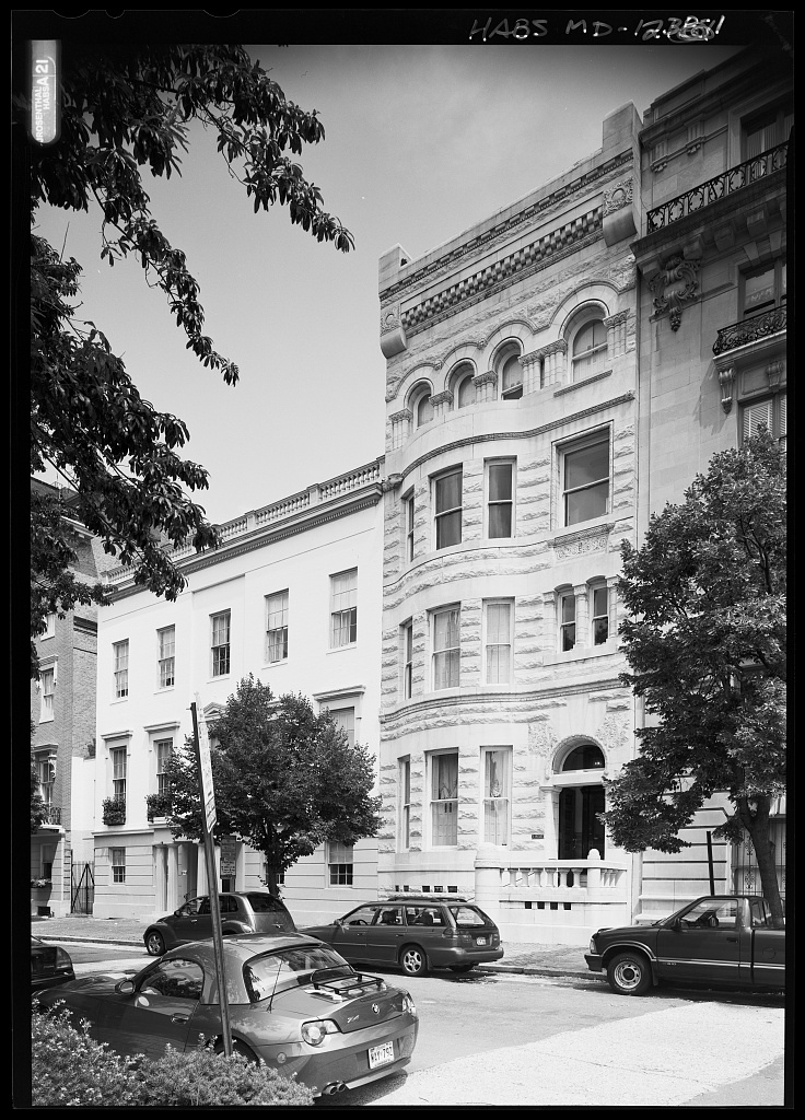 A black and white photo which shows 4 buildings, which are of different textures and heights. There are cars and trees in front of the buildings.