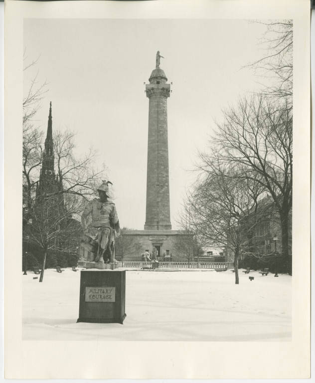 An old black and white photo shows a statue in the front, which shows a man sitting with one hand on his knee, and the other holding a sword. He is wearing a cap with a winged creature on it. He is standing on a sign that reads, “Military Courage.” Behind him is a long tower with George Washington on top. Washington is facing to the right and pointing downwards. The trees have no leaves, and there is snow on the ground.