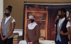 3 young teen girls perform a scene and wear costumes from the 1800s/1900s: one is white and two are Black. The two Black girls are dressed as men from the era, and the white girl a woman