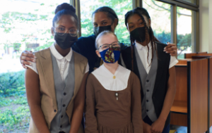 3 young teen girls wear costumes from the 1800s/1900s: one is white and two are Black. The two Black girls are dressed like men from that era and the white girl is dressed like a woman from the era. They pose with one of the girls’ moms, who is a Black woman