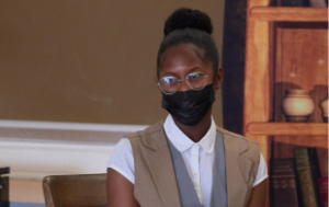 A Black teenage girl sitting in a classroom where she performs. She wears the costume of an 1800s/1900s man. There is an 1800s/1900s set behind her.