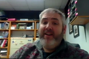 Garrett Lakes Arts Festival Board Chairman Andrew Harvey in a video screen shot. We she his face, gray hair, gray beard and mustache. He is wearing a maroon and gray sweater over a black t-shirt. In the background, we see file cabinets and supplies.