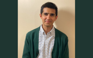 A professional headshot of photo of Aditya Desai, an Indian man with short brown hair. He wears a pine green cardigan and a white button-down shirt with a pattern on it (that includes a matching green, blue, and light pink.)