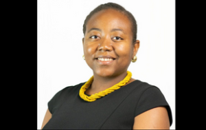 A professional headshot of Alexandra Cenatus, a Black woman with short hair. She wears a dressy black top, a yellow beaded necklace, and gold earrings. The background of the picture is white.