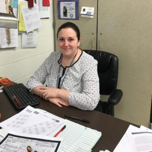 Amie Dryer, a white woman, sits at her desk. She wears her brown hair pulled back and a white blouse with black trim and black polka dots. To her left is her bulletin board and she sits in front of her file cabinet.