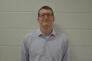 A white man with glasses wears a light-colored button-down shirt, stands in front of a white wall.