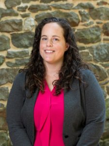 A white woman with brown curly hair stands in front of a brick background. She wears a pink button-down shirt and dark gray cardigan.