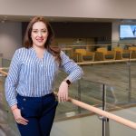 Alejandra Balcázarzar, a young to middle-aged light-skinned Latine woman, in a professional or classroom building on a college campus. She smiles and has long brown hair, wears a navy and light blue striped blouse, and navy slacks.