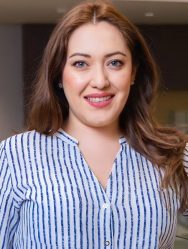 Alejandra Balcázarzar, a young or middle-aged light-skinned Latine woman, in a professional or classroom building on a college campus. She smiles and has long brown hair, wears a navy and light blue striped blouse, and navy slacks.