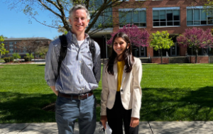 Devin Page, a middle-aged white male, stands next to his student, Zara Ahmed. They stand outside of a school building. Page wears a blue patterned button-down shirt and jeans and Ahmad, with long black hair, wears a white blazer, a yellow shirt, and black pants.