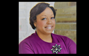 is a professional headshot of Kendra Brown. She is a Black woman with short black hair. She smiles and wears a magenta jacket and a broach. The right side of her looks to be in front of a brick wall, the left, in front of some sort of gray background