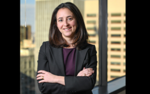 A corporate photograph of Mollie Caplis, a white woman under 50. She smiles and wears a gray blazer and a plum-colored blouse and a subtle necklace. She smiles, crosses her arms, and stands in front of an office window.