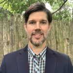 Dr. Brian Casemore, a middle-aged white man, stands outside in front of a fence with trees in the backyard and smiles. He wears a checked button-down shirt and a navy blue blazer.