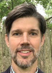 Dr. Brian Casemore, a middle-aged white man, stands outside in front of a fence with trees in the backyard and smiles. He wears a checked button-down shirt and a navy blue blazer.