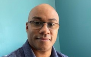 Kenneth Childs, a Black man wearing a light blue polo and glasses, sits in front of a turquoise wall. The image is from his shoulders upward.