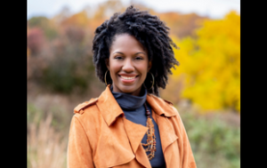 A professional photograph of Tahira Christmon, a Black woman with kinky curls. She smiles and wears a rust orange coat and dark turtleneck, gold hoop earrings and a statement necklace. She stands outside in front of trees with autumn-colored leaves and smiles.