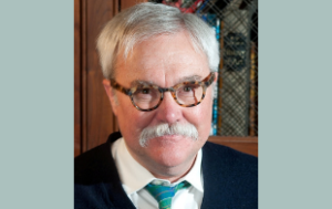 Bill Peak, a white man with thick tortoise-shell classes, white hair, and a white mustache. We see him from a little below the shoulders upwards: he wears a navy sweater, white collared shirt, and a turquoise tie.