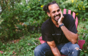 Ross Gay, a Black man with a bun, smiles. He sits on an Adirondack chair in front of some yellow flowers with lots of greenery: bushes and grass. He wears a black t-shirt and jeans.