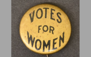 Image of a button that reads "Votes for Women." The button is yllow and the text is black.