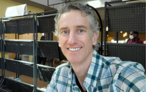 Devin Page, a white man with gray and curly hair and blue eyes smiles at the camera. He wears a blue and green shirt that is short-sleeved. He sits with his hand on his hip. Devin sits in front of what look like bins of school supplies.