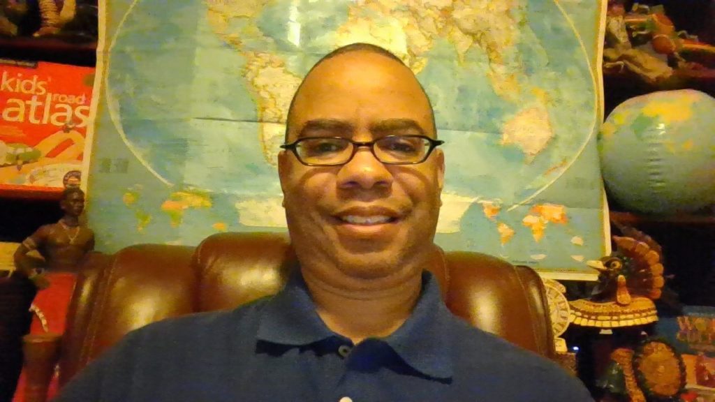 Dr. Jermaine Ellerbe, a lighter-skinned Black man, with glasses and a closely-shaved hair cut, wears a navy blue polo. He sits on a leather chair in front of a world map. Next to the map on the right, is a blow-up globe. On the map's left, is a red box that says "Kids' Road Atlas."