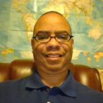 Dr. Jermaine Ellerbe, a lighter-skinned Black man, with glasses and a closely-shaved hair cut, wears a navy blue polo. He sits on a leather chair in front of a world map. Next to the map on the right, is a blow-up globe. On the map's left, is a red box that says "Kids' Road Atlas."