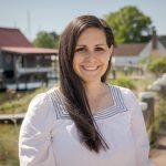 Jill Ferris, a young to middle-aged white woman with long brown hair. She smiles and wears a white peasant-style blouse with navy blue trim around the collar. She stands in front of a landscape on Maryland’s Eastern Shore, with houses, water, grass, and trees in the background.