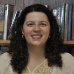 Dr. Kara French, young to middle-aged white woman with dark red or brown curly hair, smiles in front of her bookshelf. She wears a cream-colored blouse with embroidery on top, and hoop earrings.