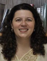Dr. Kara French, a young or middle-aged white woman with dark red or brown curly hair, smiles in front of her bookshelf. She wears a cream-colored blouse with embroidery on top, and hoop earrings.