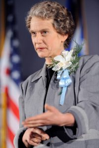 A woman with gray jack and gray wig gestures in front of a large American flag.