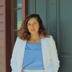 Veronica Gallardo, a Latine woman with curly brown hair, poses by standing in front of her green door to a brown house. She subtly smiles and wears a white suit and light blue blouse.