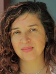 Veronica Gallardo, a young or middle-aged Latine woman with curly brown hair, poses by standing in front of her green door to a brown house. She subtly smiles and wears a white suit and light blue blouse.