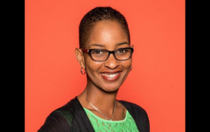 A professional headshot of Rachael Gibson, a Black woman with cropped hair and thick rimmed glasses. She smiles and wears a light green sleeveless shirt and sheer black cardigan. The background of the photo is a solid orange photo backdrop.