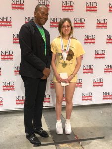 A blonde teenage girl, wearing a shirt that says "Maryland History Day," smiling after she won a Gold Medal which is placed around her neck. Next to her is a middle-aged or older Black man who wears a black suit and t-shirt. The background is a step-and-repeat that says "NHD National History Day."