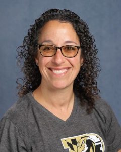 Stephanie Gomer, a white woman with dark curly hair worn down and glasses. She wears a gray t-shirt with a "P" on it, showing a falcon mascot, the mascot for Poolesville High School where she teaches.