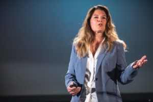 A white woman speaking in front of a blue background. She has very light brown hair and wears a white button-down shirt and a light blue blazer.