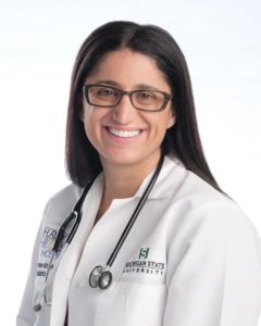 A portrait of Dr. Mona Hanna-Attisha. She is smiling, 42 and British, of Iraqi descent. She has long black hair and thick, rectangular glasses. She wears her white doctor's coat and a stethoscope.