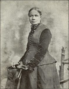 An 1893 Black and white photograph of Frances Ellen Watkins Harper, a Black woman with her hair in a bun and wearing a dark top and skirt.