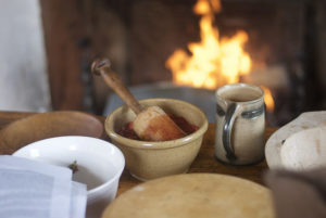 A tan, ceramic mortar and wooden pestle on a wooden table surrounded by other bowls. The table is in front of a fireplace with a fire.