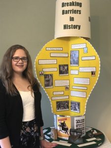 A brunette, white teenager with brown hair and glasses in a black sweater, white tank top, and black patterned skirt. She is next to her project. It is made out of yellow poster board with text and images. The title of the poster says "Breaking Barriers in History."