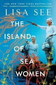 The book cover of THE ISLE OF SEA WOMEN by Lisa See. The cover is aqua with white font. Two Korean women stand in clothes, swimcaps, and goggles. They are carrying nets. They stand on a rock near the water. We also see plants.