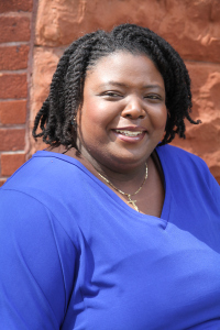 A contemporary photograph of Ida Jones, Ph.D. She is a Black woman in a royal blue sweater. She is smiling and in front of a brick background.