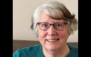 Selfie of Jacquelyn Lucy, a retired white woman smiling. She has short gray hair, glasses, and wears a teal sweatshirt.
