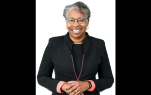 A professional headshot of Dr. Tuajuanda Jordan, an older Black woman with short gray hair and glasses. She smiles widely and wears a black suit jacket and top set. The jacket cuffs and lining are a salmon color. The background of the photo is a solid white photo backdrop.