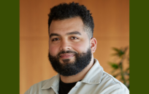 A headshot of Keith Stone sitting or standing in front of what looks like a wooden wall. Keith is a light-skinned Black man with curly black hair and a black beard. He wears a khaki-colored button-down shirt over a black t-shirt. There is a plant to his left.