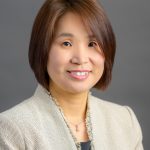 Ruth Kim, a young to middle-aged Korean American woman, smiles. She wears a dressy tan jacket, dark grey top, and drop necklace. The background to her headshot is a simple gray.