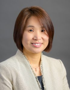Ruth Kim, a young or middle-aged Korean or Korean American woman, smiles in front of a solid gray photo backdrop. She wears a dressy tan jacket, dark grey top, and drop necklace.