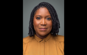 A professional headshot of Dr. Shauna Knox, a Black woman with shoulder-length locs. She smiles subtly and wears a golden-colored button-down shirt with all the buttons buttoned. 