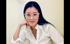 A professional photograph of Soo Koo, a young or middle-aged East Asian woman. She wears her black hair down, a white button-down shirt, and a chunky silver necklace, and small hoop earrings. She has large, clear-rimmed, round glasses and leans her hand on the bottom half of her face in an engaged manner. The background is a light tan photo backdrop. 